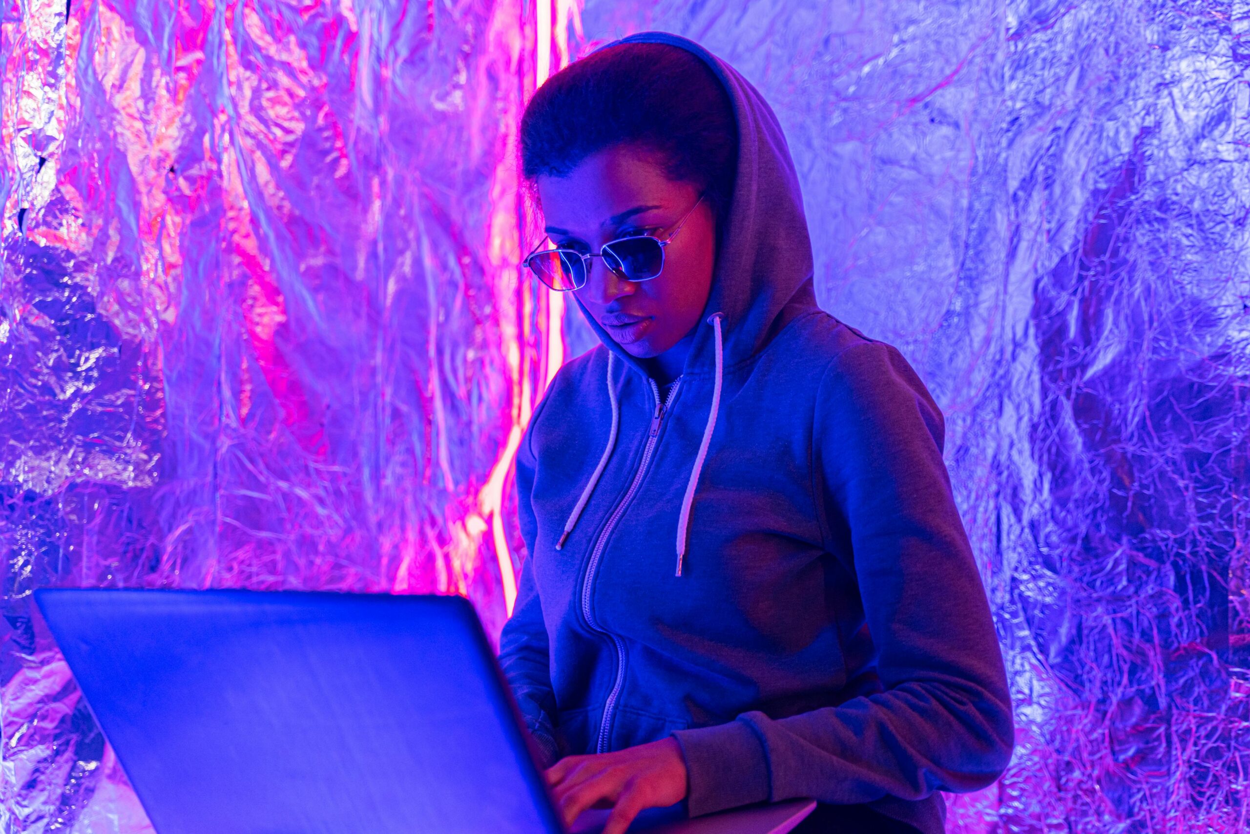 A woman in a hoodie works on a laptop in a futuristic neon-lit space.